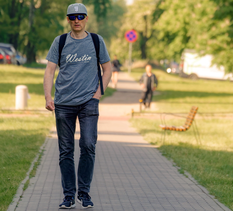 28 - An individual walking along a paved park path while wearing casual apparel and carrying a backpack